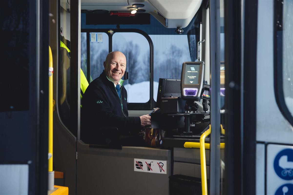 Bilde av smilende bussjåfør sittende i førersetet på bussen. Foto. - Klikk for stort bilde