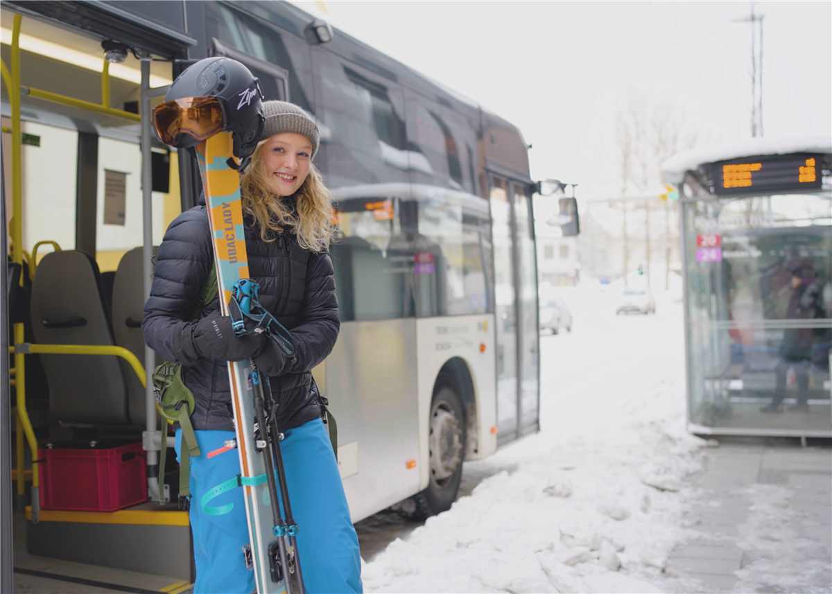 Smilende jente med skiutstyr og vintertøy foran buss i sentrum. Foto. - Klikk for stort bilde